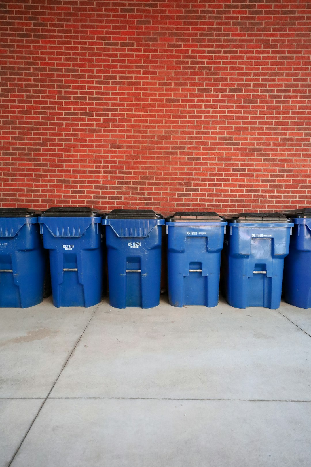 a-row-of-blue-trash-cans-sitting-in-front-of-a-brick-wall-udhrcrnow7w