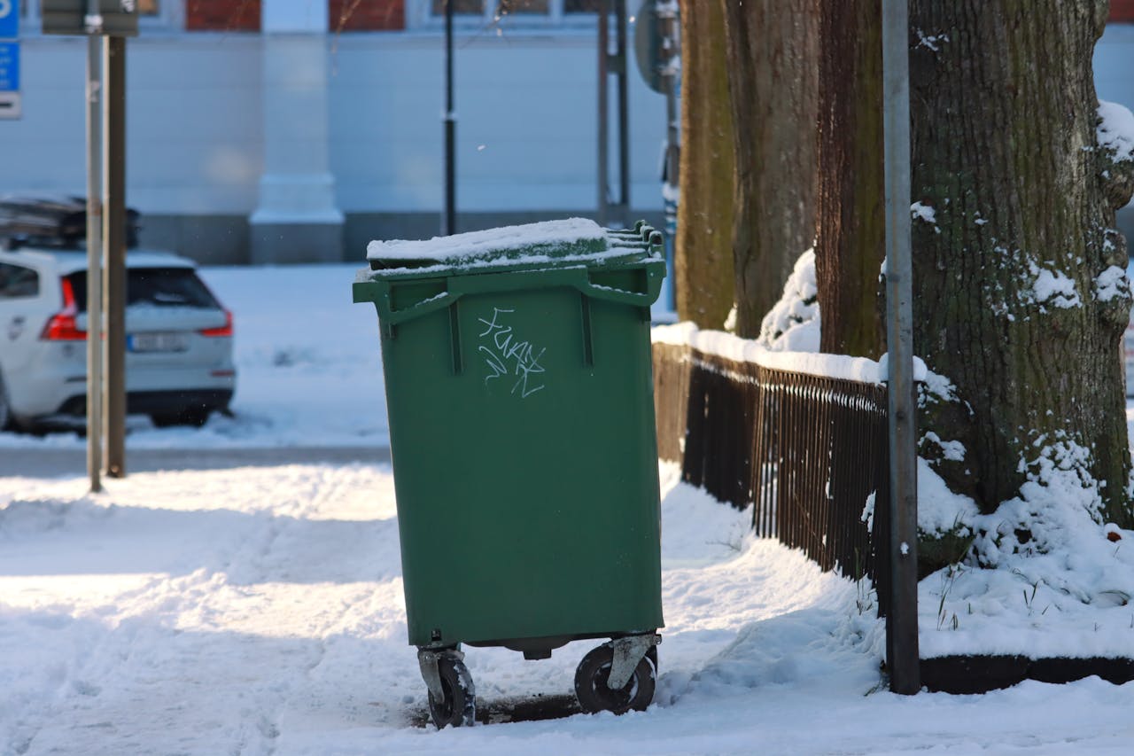 A green trash bin covered in snow on a suburban street during winter.
