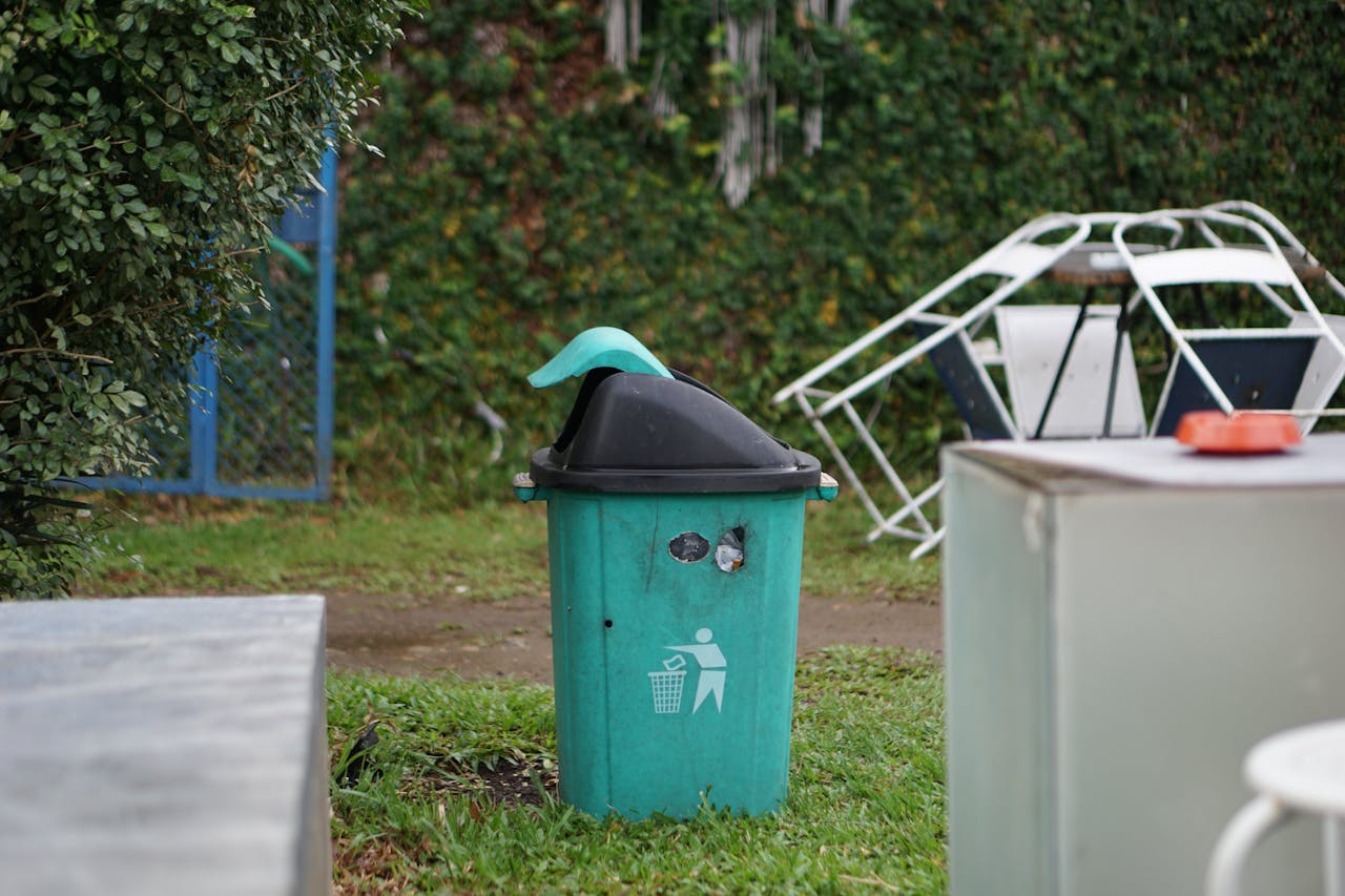 A green trash bin in a grassy outdoor area in Palembang, Indonesia.